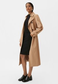 Beiger Trenchcoat aus glattem Stoff, mit einem doppelreihigen Design, breiten Revers und taillierten Ärmeln. Kombiniert mit einem schwarzen Kleid und Schuhen.