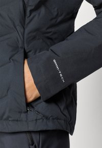 Temno izolirano jopico z gladko teksturo. Ima zadrgo, nastavljiv rob in logo "OMNI-TECH" na manšeti.