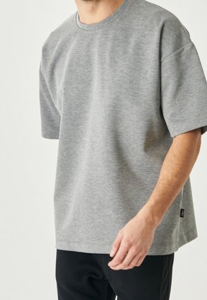 Man wearing a loose, light grey short-sleeve crewneck T-shirt and black pants, standing against a plain light background.