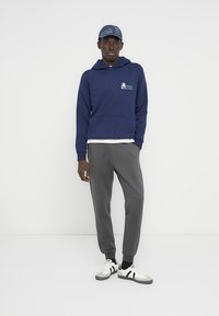 Φούτερ με κουκούλα σε ναυτικό μπλε χρώμα με μπροστινή τσέπη, συνδυασμένο με γκρι στενά παντελόνια φόρμας και λευκά αθλητικά παπούτσια με μαύρες λεπτομέρειες.