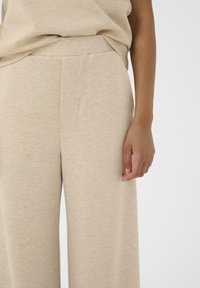 Beigeweite Hose aus weichem Strickstoff, mit einem flachen Bund und seitlichen Eingrifftaschen, glatte Oberfläche mit einem melierten Muster.