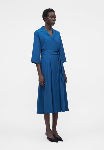 Γυναίκα που στέκεται φορώντας ένα μπλε μίντι φόρεμα με γιακά, μανίκια τριών τετάρτων, δεμένη ζώνη στη μέση και μαύρα χαμηλά μυτερά παπούτσια.