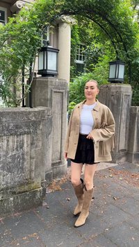 Beige Fransenjacke über einem weißen Oberteil, kombiniert mit einem schwarzen, gerafften Rock und braunen Stiefeln bis zum Knie. Steinmauer und Grünpflanzen im Hintergrund.