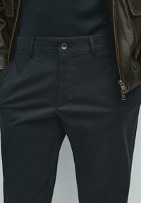 Zwarte broek met een knoopsluiting, twee voorzakken en een gladde, licht getextureerde stof. Bevat riemlussen en heeft een getailleerde pasvorm.