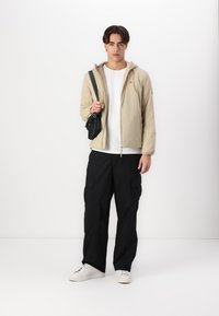 Beige lett jakke med hette over en hvit t-skjorte. Svart cargo-bukser og hvite sneakers. Liten svart veske slengt over skulderen.
