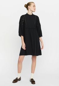 Schwarzes Kleid mit einem gefalteten Vorderteil, Spitzenverzierungen und Ballonärmeln. Der Stoff wirkt weich, mit einem lockeren Schnitt, der über die Knie fällt.