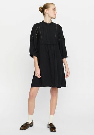 Vestito nero con una parte frontale plissettata, dettagli in pizzo e maniche a palloncino. Il tessuto sembra morbido, con una vestibilità ampia che scende sopra le ginocchia.
