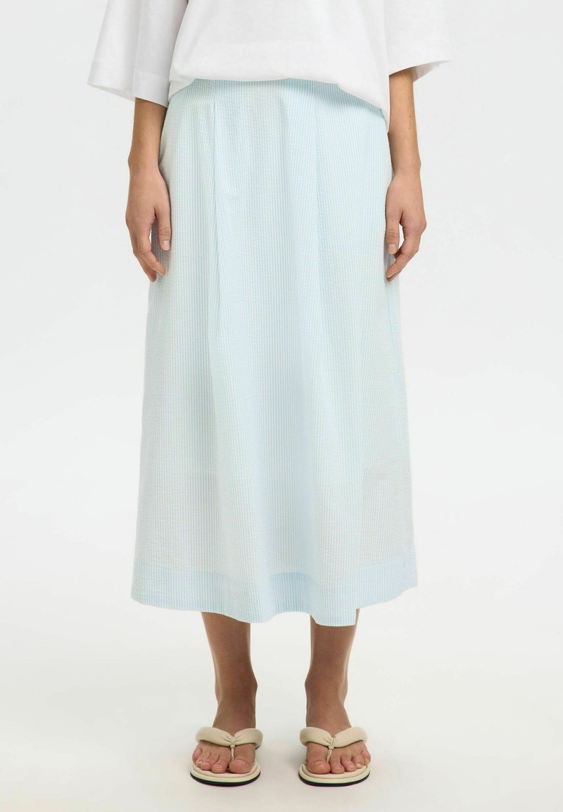 Personne portant une jupe midi rayée bleu clair et blanc, un haut blanc et des sandales beige contre un fond blanc uni.
