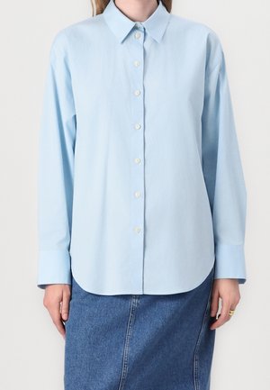 Lyseblå langærmet skjorte med knapper, båret over en blå denims nederdel, med synlig krave og manchetter, og hænder hvilende ved siderne.