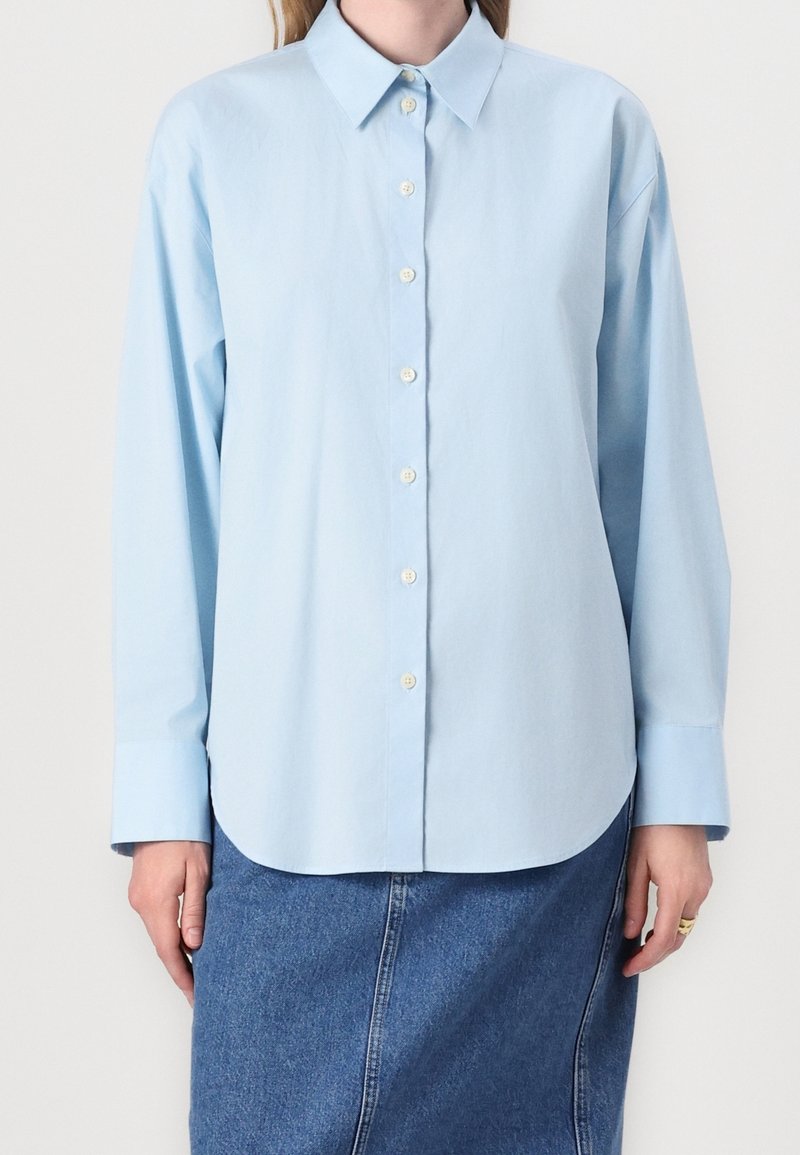 Chemise bleu clair à manches longues boutonnée portée sur une jupe en jean bleu, avec col et poignets visibles, les mains reposant sur les côtés.