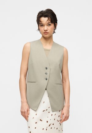 Femme portant un blazer beige sans manches à trois boutons par-dessus un haut clair et une jupe blanche à pois marron, debout devant un fond uni.