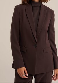 Kobieta w dopasowanej, ciemnobrązowej marynarce zapinanej na jeden guzik, z kieszeniami na klapki i pasującymi spodniami, z ręką ozdobioną srebrnym pierścionkiem.