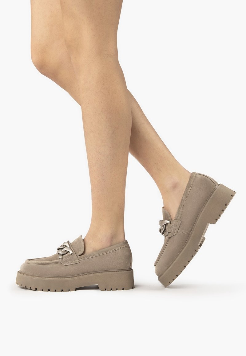 Jambes portant des mocassins à plateforme en daim beige avec des semelles épaisses à crampons et un détail chaîne en métal sur le dessus, sur un fond blanc.