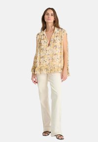 Blusa floral amarilla y crema con un escote en V profundo, dobladillo con volantes y mangas cortas, combinada con pantalones blancos de campana y sandalias negras.