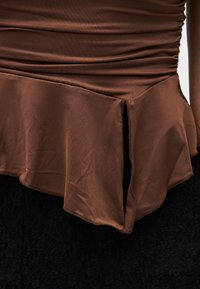Gros plan sur un haut marron froncé avec un ourlet asymétrique et une fente frontale, porté par-dessus une jupe ou un pantalon noir texturé.
