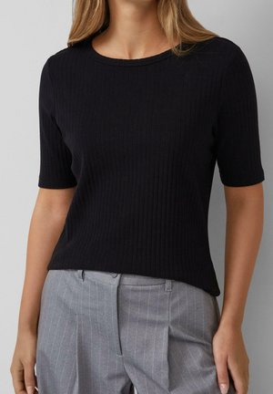 Femme portant un top côtelé noir moulant à manches courtes et un pantalon gris à rayures taille haute, debout les bras détendus.