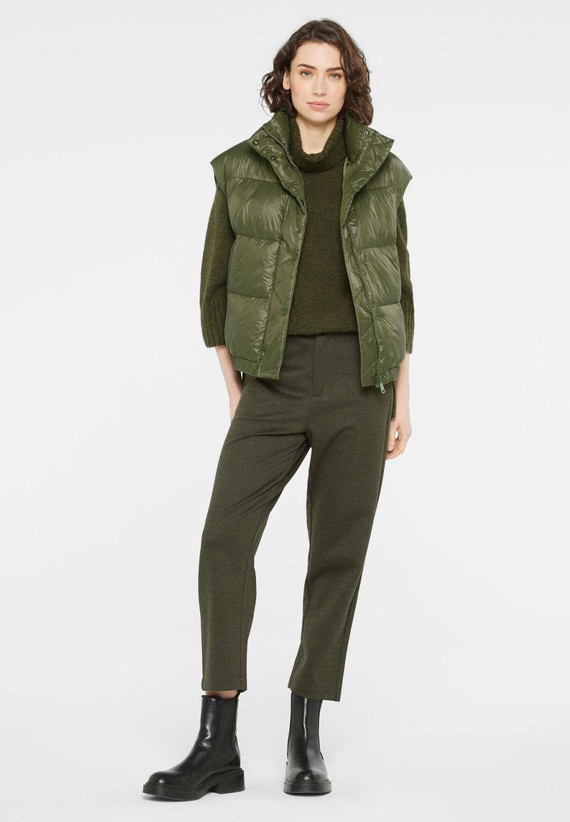 Grüne gepolsterte Weste mit hohem Kragen, getragen über einem dunkelgrünen Pullover. Kombiniert mit strukturierten grünen Hosen und schwarzen Ankle Boots.