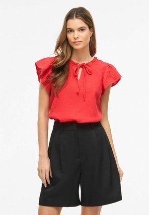 Femme aux longs cheveux bruns portant un chemisier rouge à manches courtes avec un détail noué et un short noir taille haute arrivant au genou.