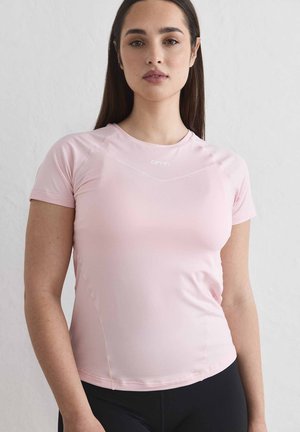 Jonge vrouw met lang donker haar, gekleed in een lichtroze nauwsluitend shirt met korte mouwen en een zwarte broek, staand tegen een effen lichte achtergrond.