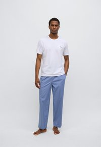 Maglietta bianca in cotone con logo "BOSS" nero sul petto, abbinata a pantaloni azzurri, larghi e comodi. Modello in piedi a piedi nudi.