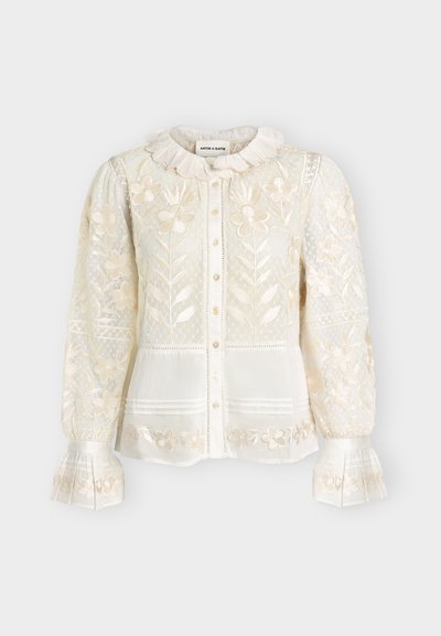 Blouse blanche manches longues avec broderies florales, tissu transparent à motifs, col et poignets à volants, fermeture boutonnée devant et délicats détails en dentelle.