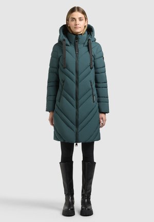 Donna in piedi che indossa un cappotto trapuntato color teal lunghezza ginocchio con cappuccio, leggings neri e stivali neri alti al ginocchio su uno sfondo semplice.