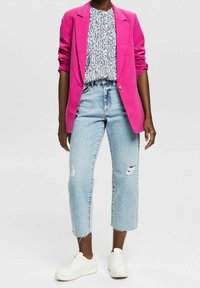 Blazer rose vif porté sur un haut à motifs, associé à un jean bleu clair déchiré et des baskets blanches. Coupe décontractée avec jambe raccourcie.