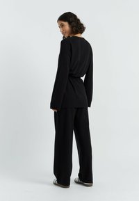 Zwarte gebreide top met lange mouwen, relaxte pasvorm en een ceintuur in de taille. Gecombineerd met zwarte brede broeken. Glad textuur en minimaal ontwerp.