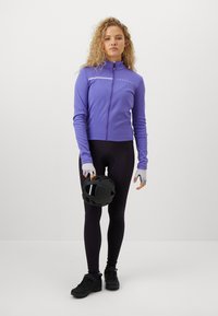 Kobieta stojąca w stroju rowerowym, trzymająca czarny kask, ubrana w fioletową kurtkę na zamek, czarne legginsy, rękawiczki i czarne buty na tle białym.