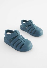 Blå gummitofflor med stängd tå, detalj med remmar och ventilationshål, med en strukturerad yta och en robust sula.