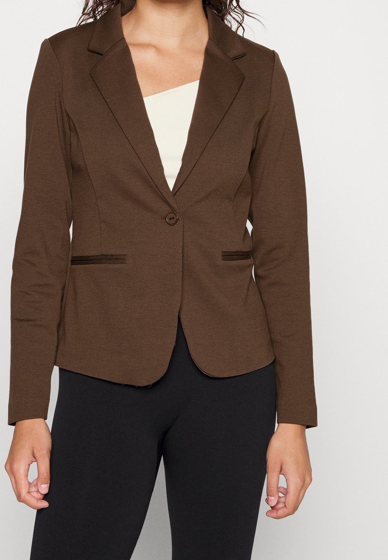 Femme portant un blazer marron ajusté à un seul bouton et un pantalon noir, debout devant un fond blanc uni.