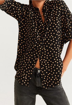 Black button-up shirt with beige polka dots worn with dark gray jeans, showing midsection and part of raised arm.