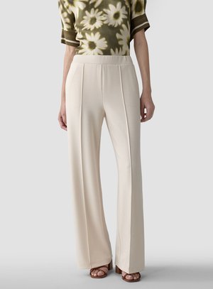 Frau trägt beige hoch taillierte, weit geschnittene Hosen, kombiniert mit braunen offenen Absätzen und einem grünen floralen Oberteil.