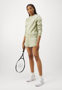 Felpa di cotone verde chiaro con pantaloni corti abbinati, con un logo discreto. Il modello tiene una racchetta da tennis, indossa scarpe da ginnastica e calzini bianchi.