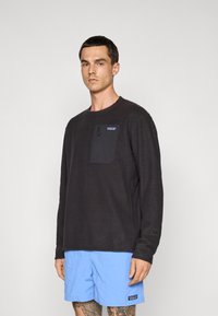 Un homme portant un pullover noir à manches longues de Patagonia avec une poche zippée sur la poitrine et un short bleu clair de Patagonia, se tenant debout contre un fond blanc.