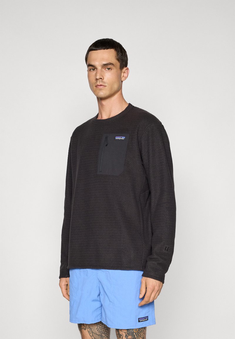 Un homme portant un pullover noir à manches longues de Patagonia avec une poche zippée sur la poitrine et un short bleu clair de Patagonia, se tenant debout contre un fond blanc.