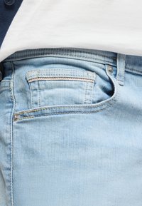 Φωτεινό μπλε τζιν με λεπτομερή ραφή στην μπροστινή τσέπη, φορεμένο με μια λευκή πουκάμισο μέσα στην μπλούζα και ένα σκούρο πουλόβερ.