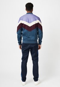 Men's athletic tracksuit comprised of a blue jacket with chevron pattern in purple, white, and burgundy; paired with navy pants.