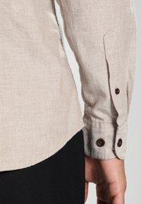 Beżowa lniana koszula z długim rękawem, z mankietami na guziki i zaokrąglonym dołem. Posiada ciemnobrązowe guziki i teksturowaną tkaninę.