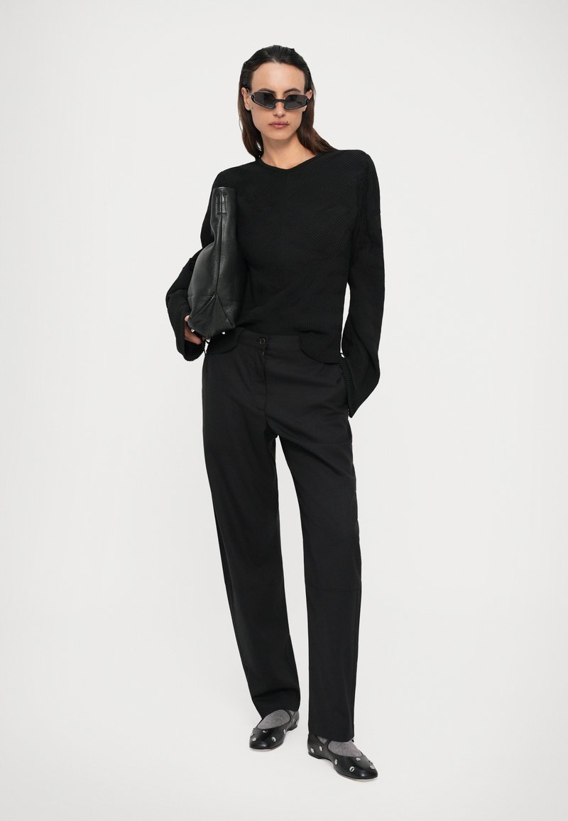 Femme portant un pull texturé noir et un pantalon, tenant un sac en cuir noir, portant des lunettes de soleil sombres et des ballerines noires avec des chaussettes grises.