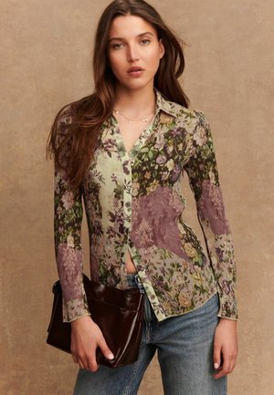 Camicia con fantasia floreale, bottoni e texture a coste in tonalità di verde e viola, abbinata a una borsa a mano marrone scuro e jeans in denim.