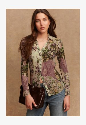 Camicia con fantasia floreale, bottoni e texture a coste in tonalità di verde e viola, abbinata a una borsa a mano marrone scuro e jeans in denim.