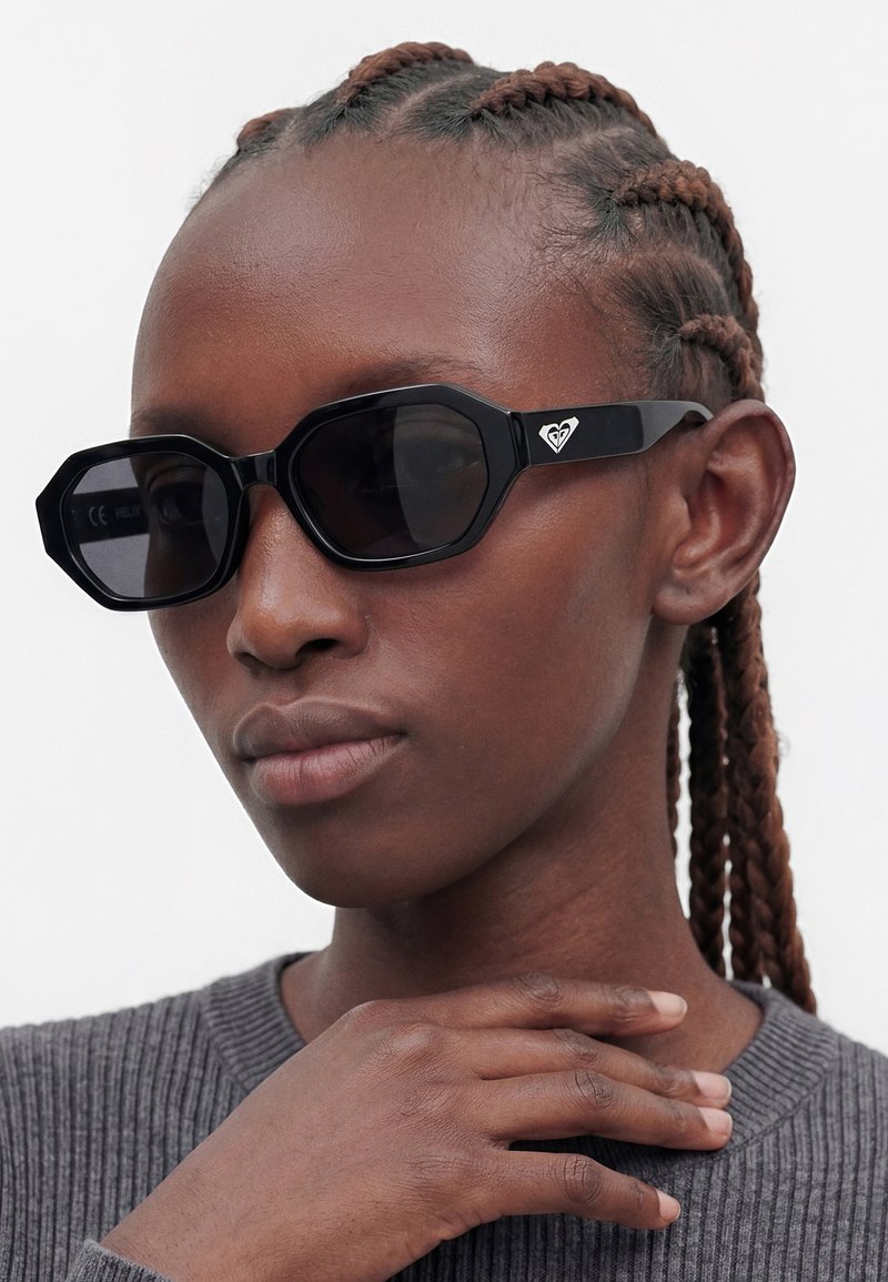 Gros plan d'une personne aux cheveux tressés portant des lunettes de soleil noires géométriques et un haut gris côtelé, main reposant délicatement sur le cou.