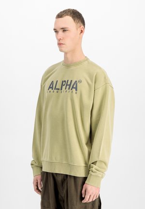 Junger Mann trägt ein hell olivgrünes Sweatshirt mit dem Schriftzug "ALPHA INDUSTRIES" und steht vor einem einfarbigen weißen Hintergrund.
