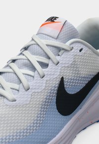 Hele sinine Nike jooksujalats võrgust pealispinnaga, tekstureeritud pinnaga, musta logoga ja hallide paeltega. Kontsal on oranž aksent.