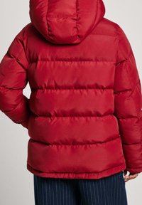 Röd puffjacka med horisontella quiltade paneler, huva och elastiska ärmmuddar, kombinerad med marinblå randiga byxor.