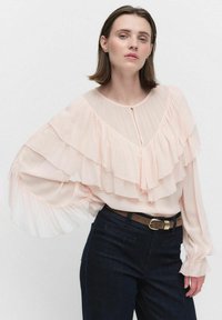 Vaaleanpunainen pusero, jossa on kerrokselliset frillat, V-pääntie ja rypytetyt hihat. Yhdistetty tummansiniset farkut ja ruskea vyö, jossa on kultainen solki.