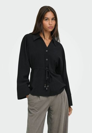 Femme portant un chemisier noir boutonné avec une ceinture à la taille et un pantalon gris à fines rayures, posant avec une main dans la poche sur un fond uni.