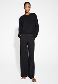 Schwarze Strickjacke mit rundem Ausschnitt und Knopfverschluss, kombiniert mit schwarzen weit geschnittenen Hosen und schwarzen Flip-Flop-Sandalen.