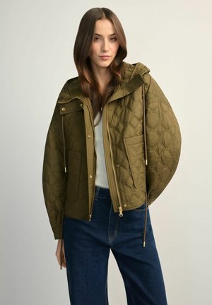 Kobieta ubrana w pikowaną oliwkowozieloną kurtkę z kapturem, zamkiem błyskawicznym, zatrzaskami, dużymi kieszeniami, w połączeniu z niebieskimi dżinsami i białą bluzką.